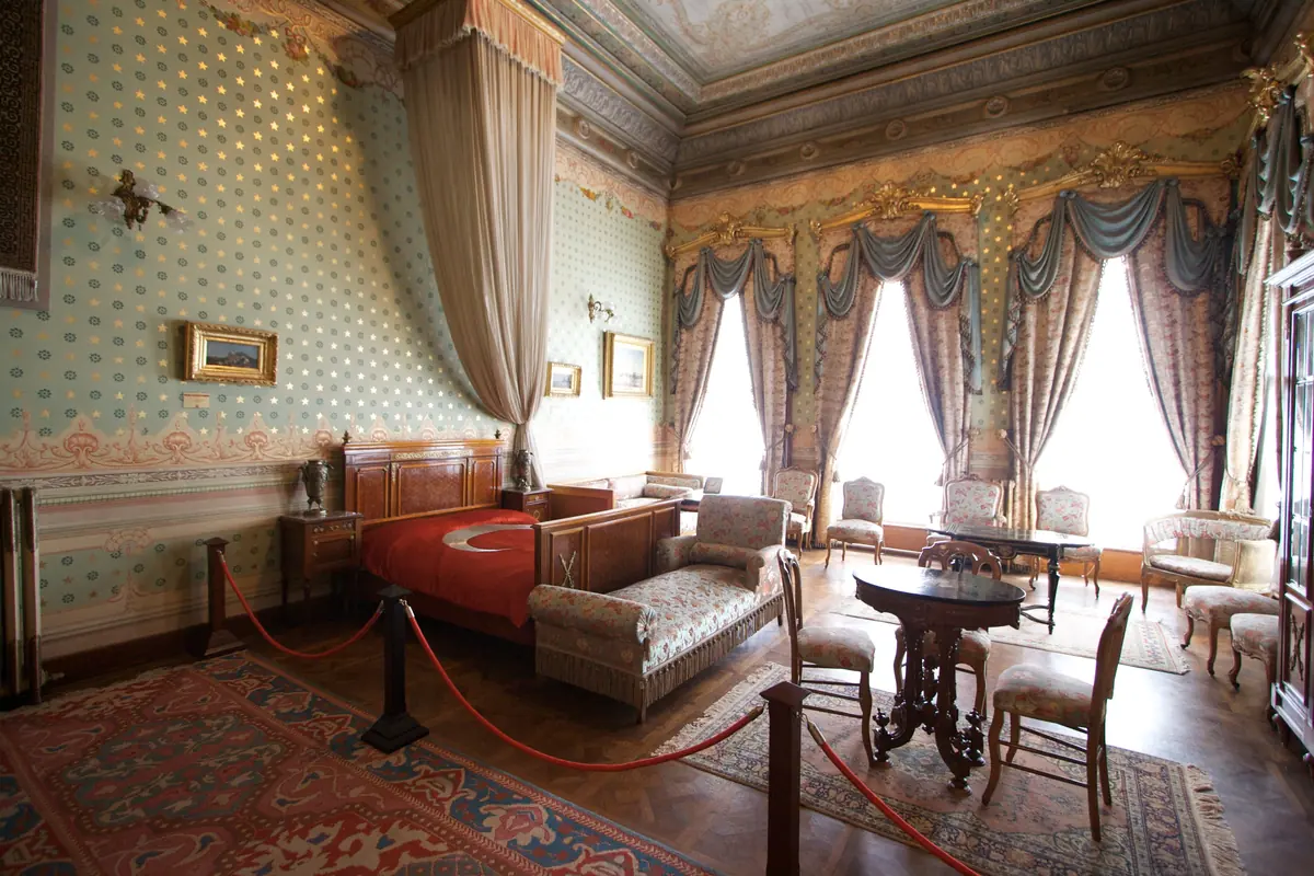 A bedroom in Dolmabahçe Palace with a bed draped in a Turkish flag, tall curtained windows, a small table and chairs, and a patterned carpet
