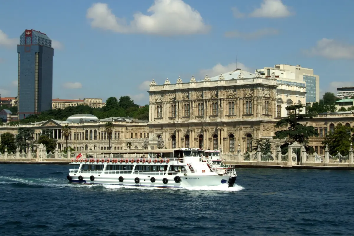 A white passenger ferry crossing the Bosphorus in front of the palace's long façade, with a tall tower block behind.