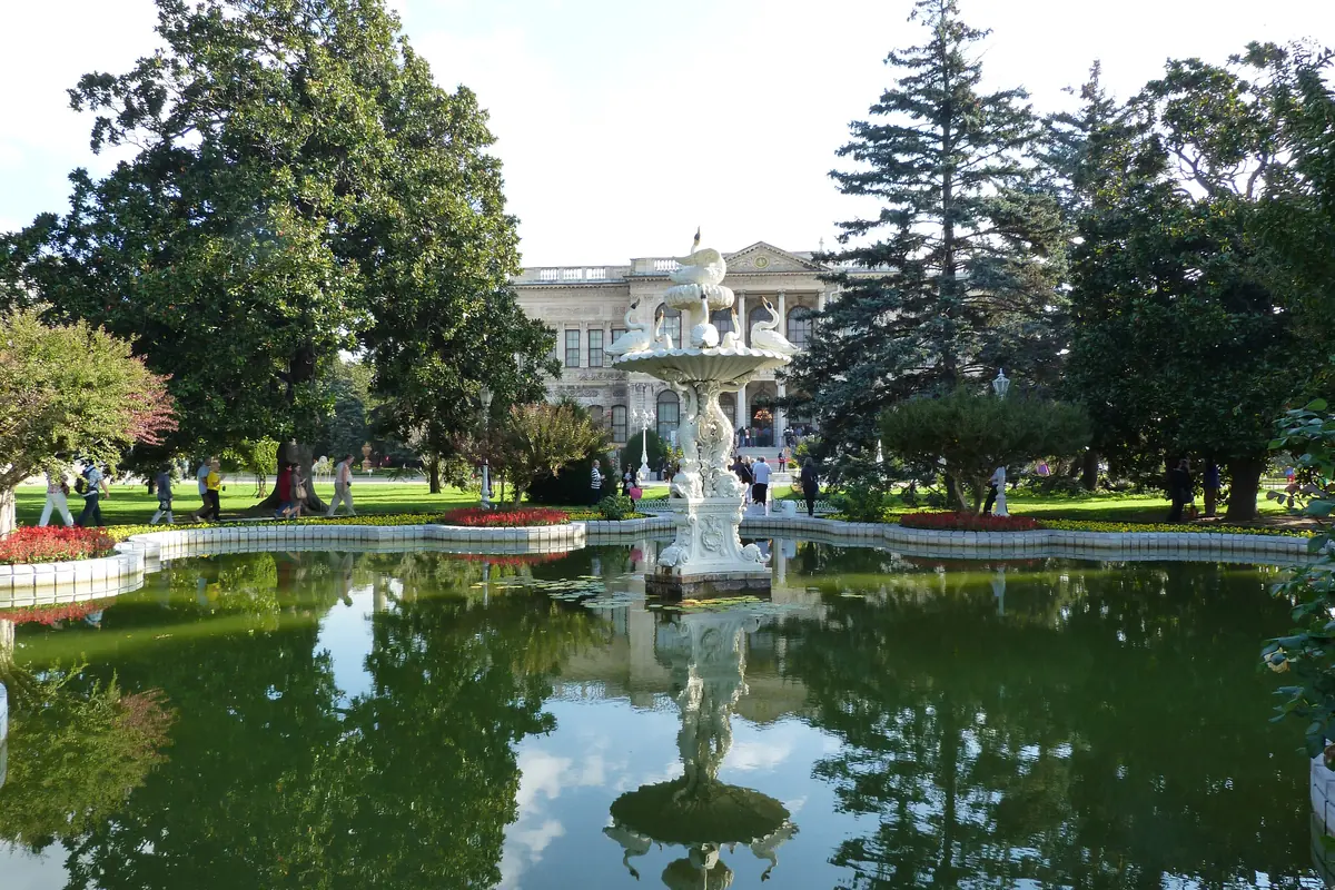 A pond with a stone fountain in the palace garden, reflecting trees, with the palace building visible beyond the lawn.