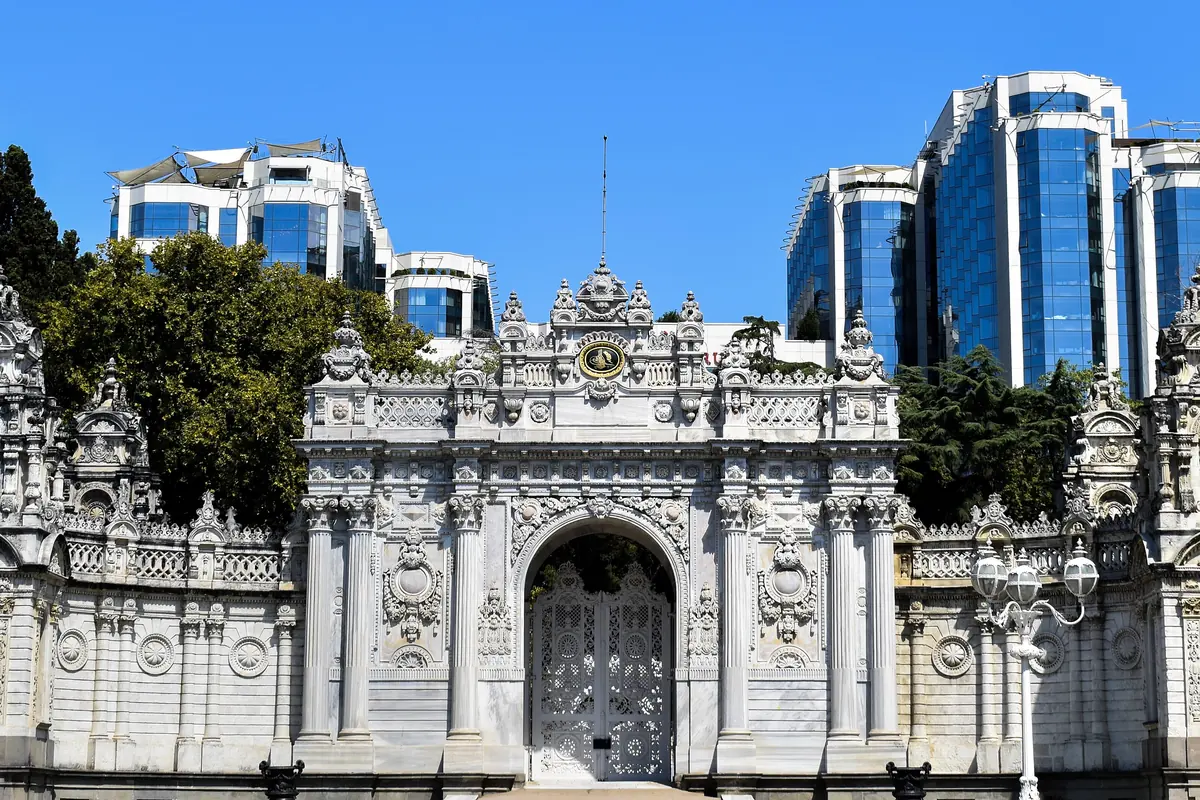The ornate white Imperial Gate of the palace under a blue sky, with glass office towers rising behind the trees.