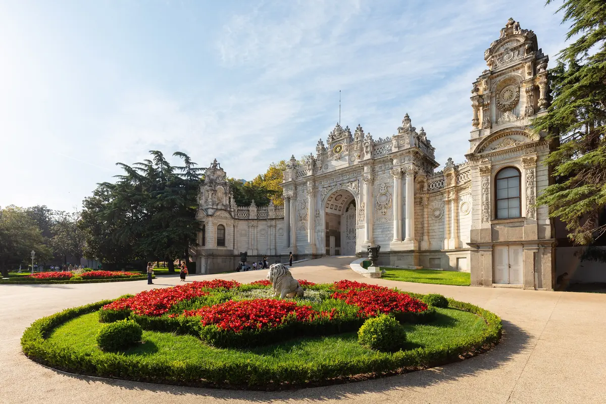 The Sultan's Gate of the palace beside a round flowerbed of red flowers on a sunny day.