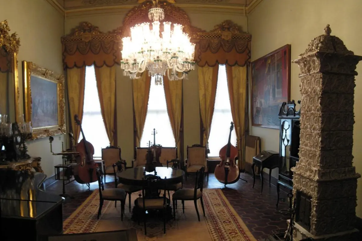 A sitting room in the Harem of Dolmabahçe Palace with a chandelier, gold curtains, a round table, double basses and a tall tiled stove
