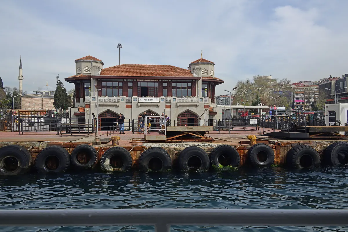 The Beşiktaş ferry pier, a red-roofed pavilion with two small clock towers, seen across the water from a boat with tyre fenders along the quay