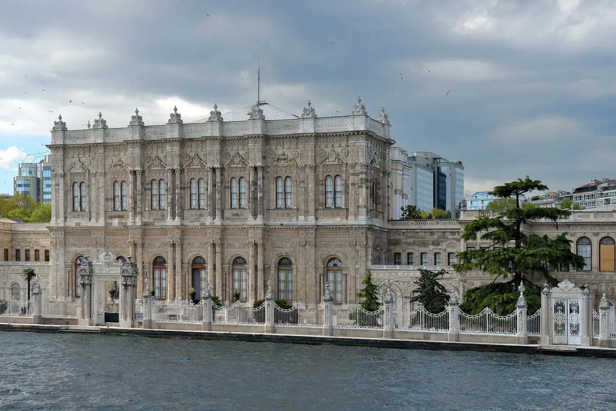 The long sea-facing façade of the palace and the white Sultan's Gate seen from the Bosphorus, with a cypress and a grey sky behind.