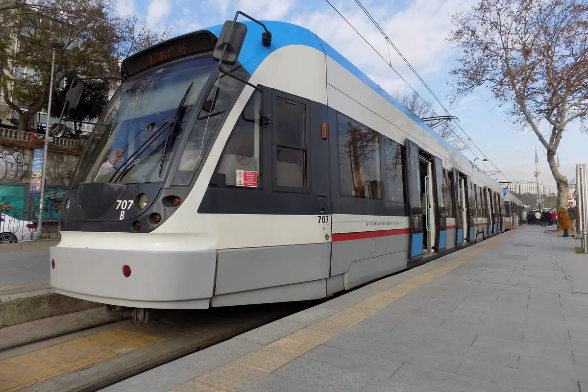 A grey and white T1 tram standing at a platform at Kabataş in Istanbul on a bright day, with bare trees beside the track